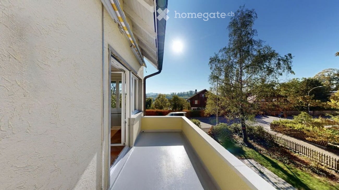 Balcony with white flooring and yellow railing, overlooking a green garden and distant houses under a clear blue sky.