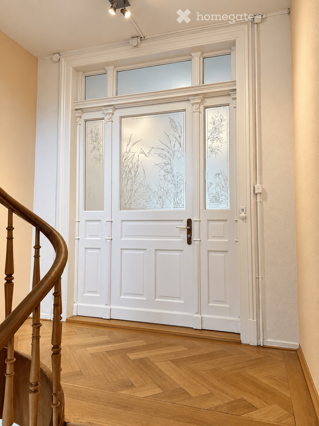 Elegant hallway with wooden flooring, white paneled doors featuring decorative glass panels, and a wooden staircase railing on the left side.