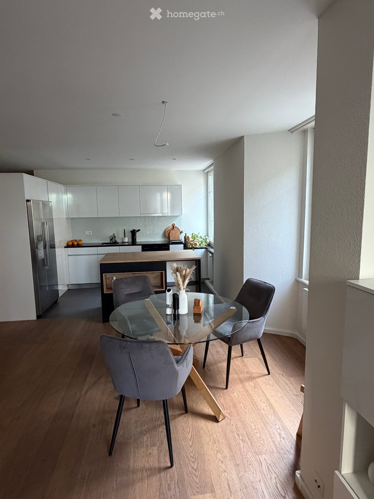 Modern dining area with a glass dining table and four grey chairs, adjacent to a sleek kitchen with white cabinets, stainless steel appliances, and a wooden kitchen island.