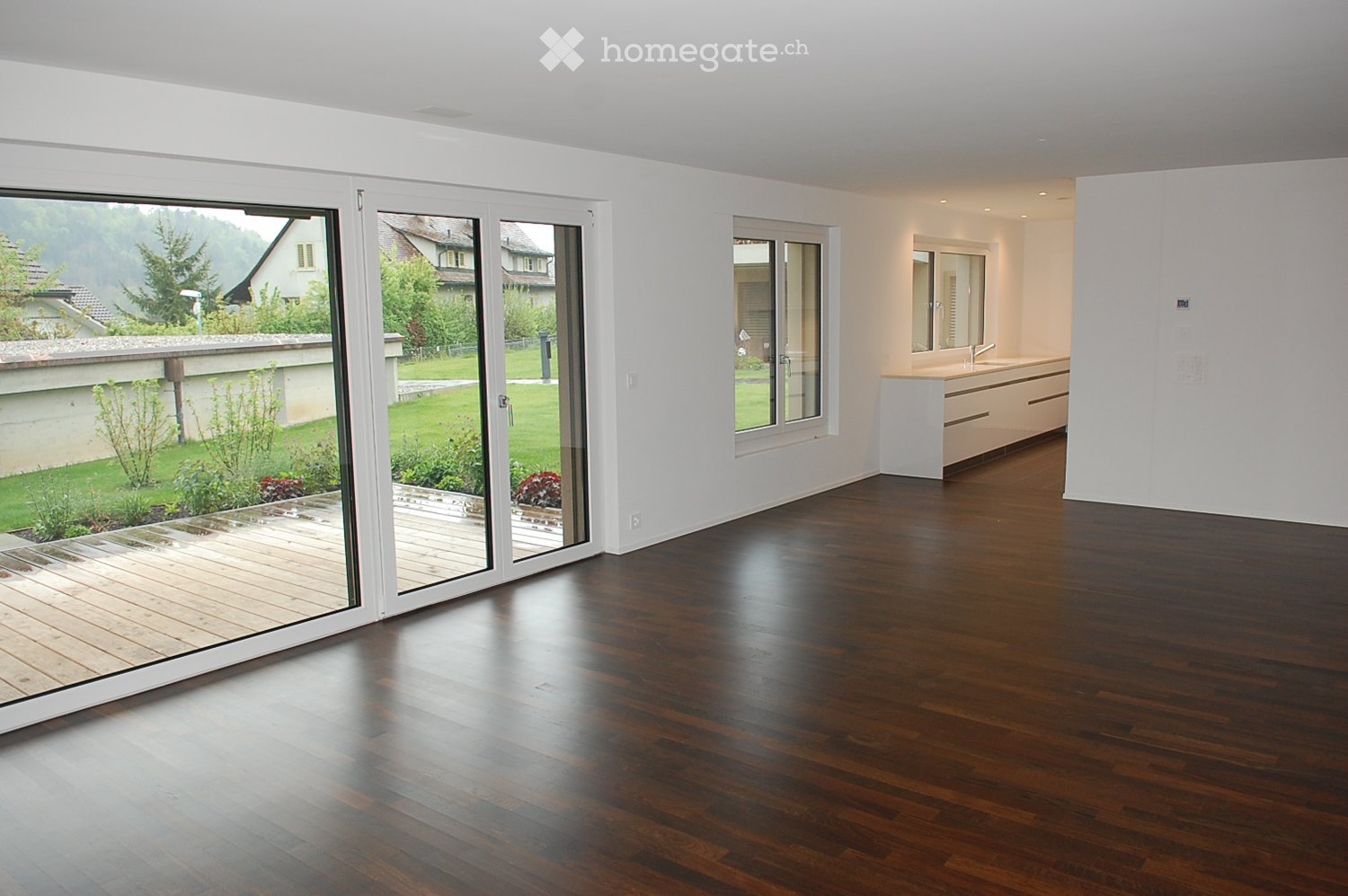 Spacious, unfurnished room with dark hardwood flooring, large sliding glass doors leading to a wooden deck, and a view of a green garden. A small, modern bathroom is visible in the background.