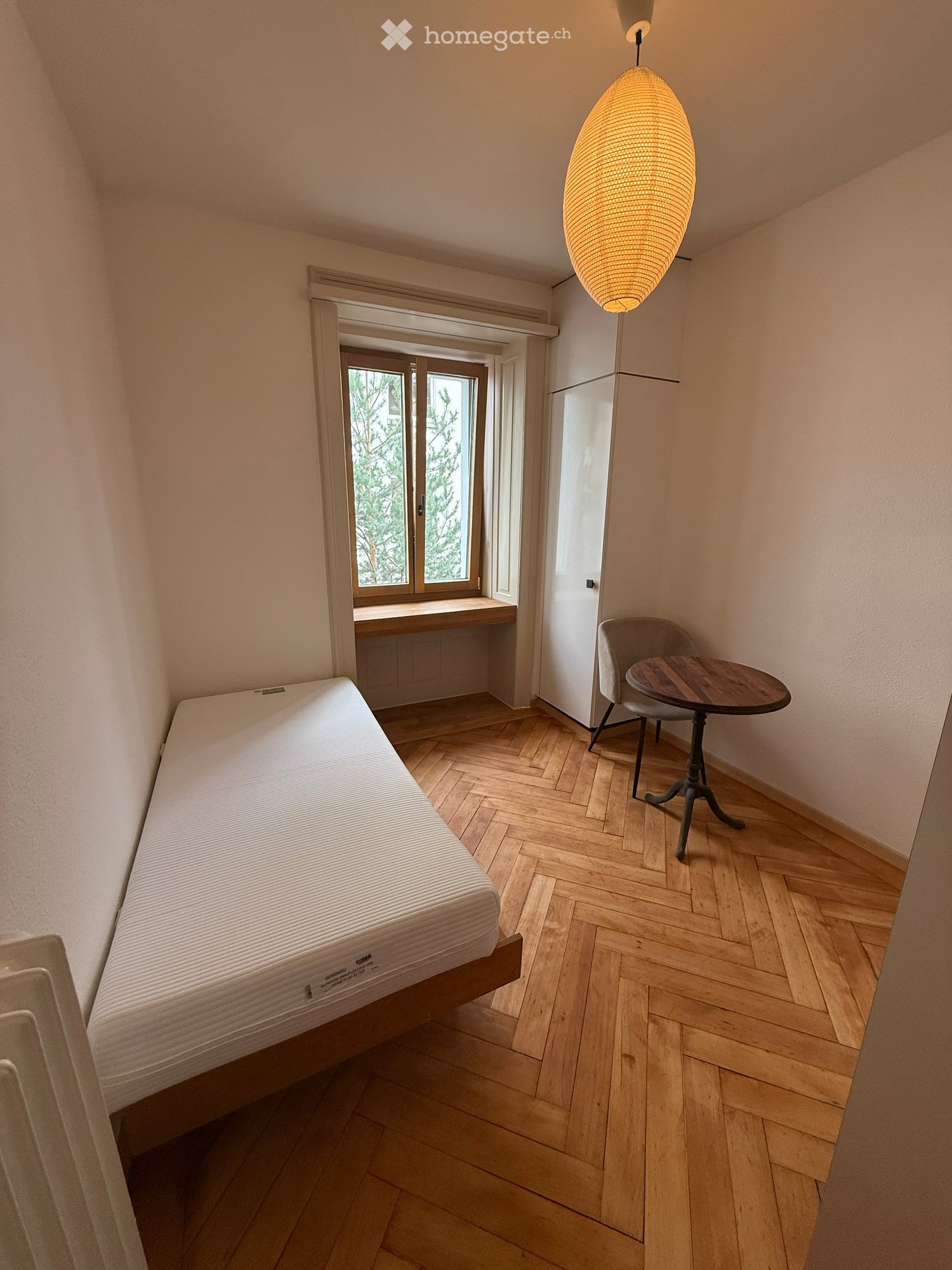 Cozy bedroom with wooden herringbone flooring, a single bed, a small round table with a chair, and a hanging lamp.