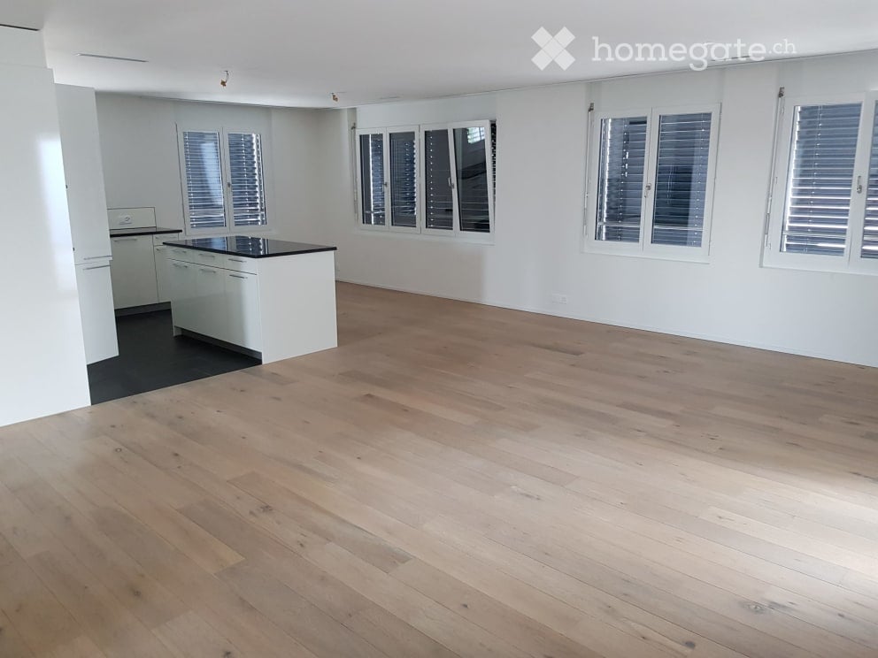 Empty room with light wooden flooring, white walls, and large windows with shutters. A small kitchen area is visible with white cabinets and a black countertop.