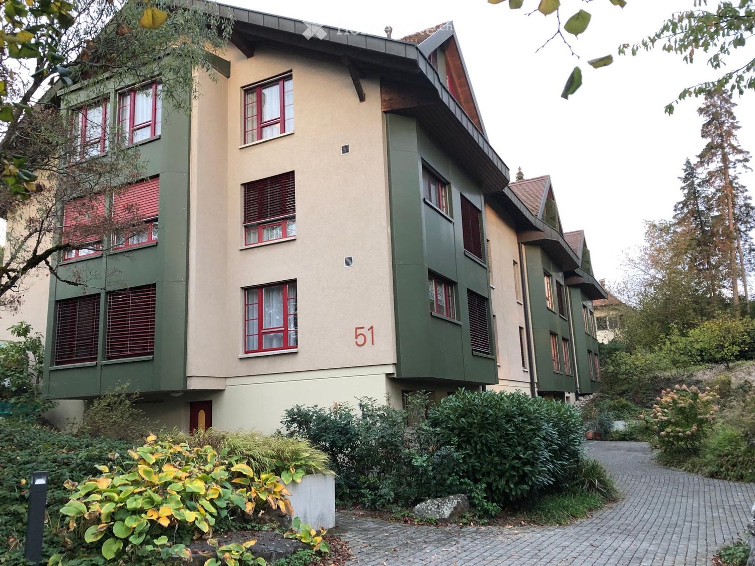 A multi-story residential building with a beige and green facade, red-framed windows, and a well-maintained garden in front.