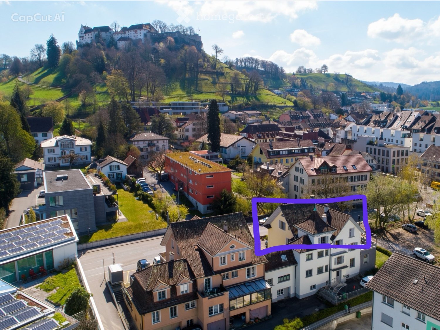 Aerial view of a town with multiple residential buildings, green spaces, and a castle on a hill in the background.