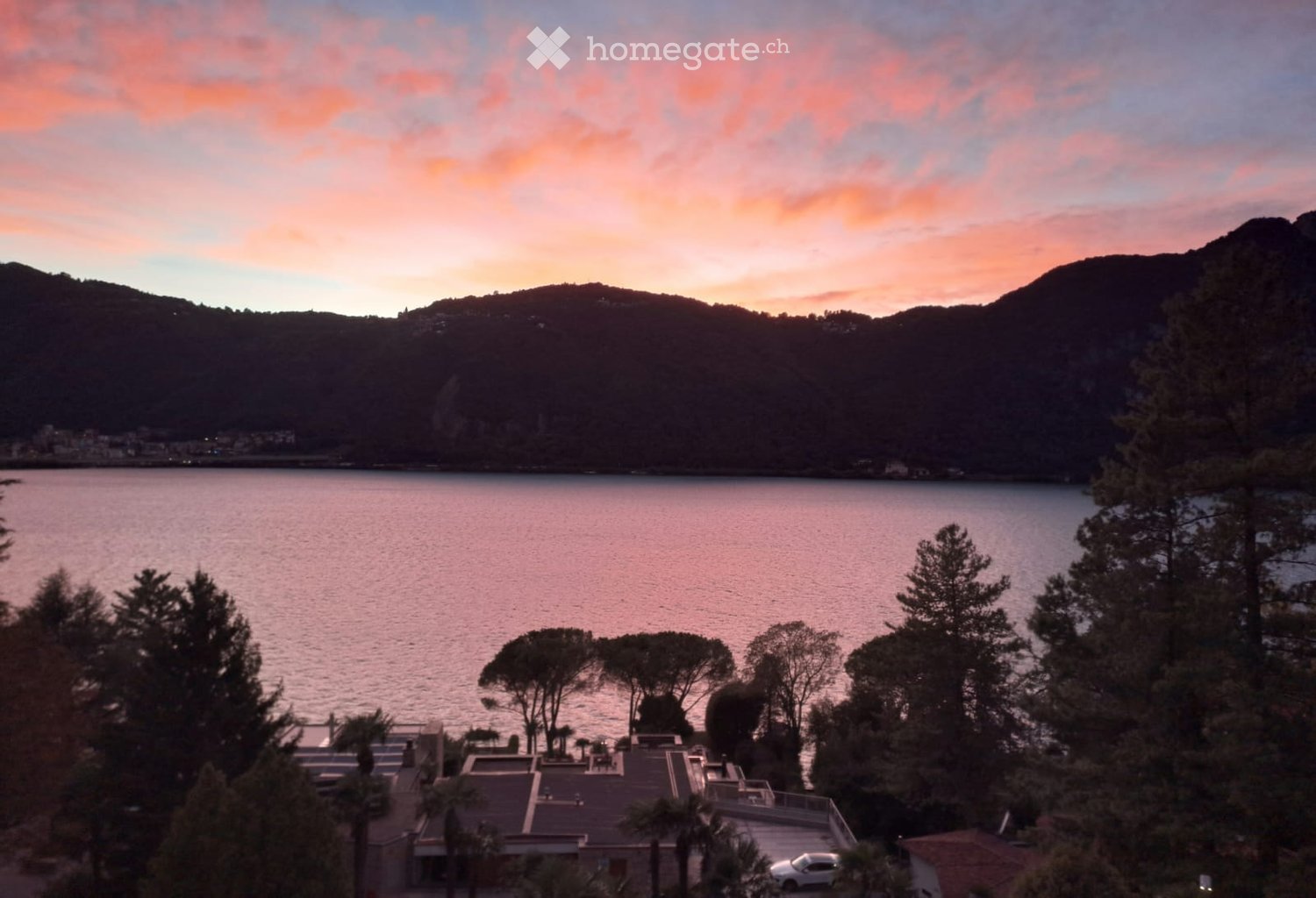 A scenic view of a lake surrounded by trees and mountains, with a house and parking area in the foreground.