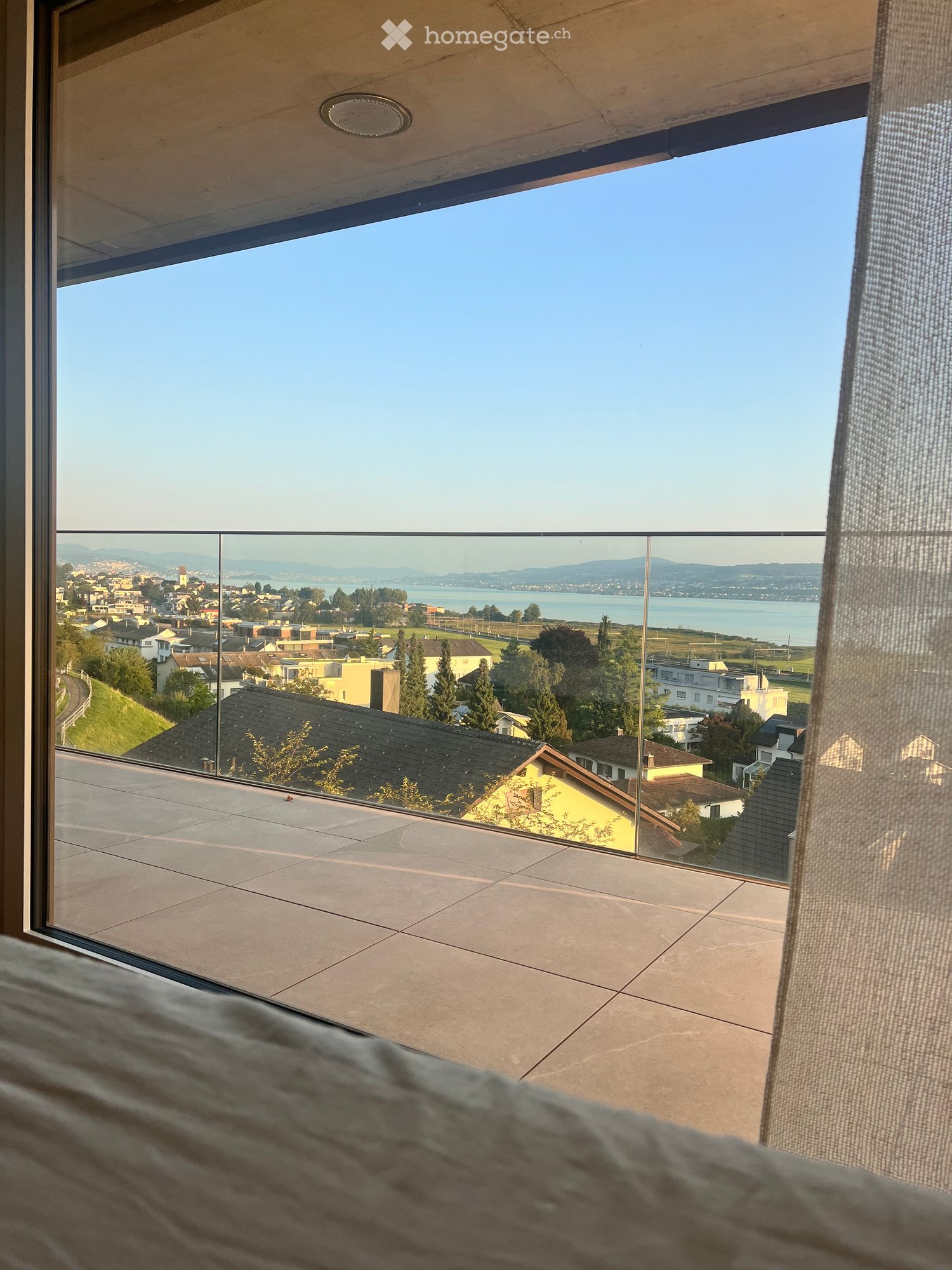Modern balcony with large glass panels, tiled floor, and scenic view of a town, water, and distant mountains.