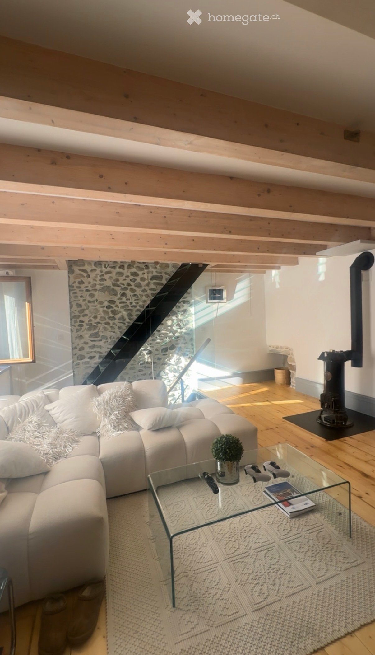 Modern living room with exposed wooden beams, a white sectional sofa, a glass coffee table, a small potted plant, and a fireplace.