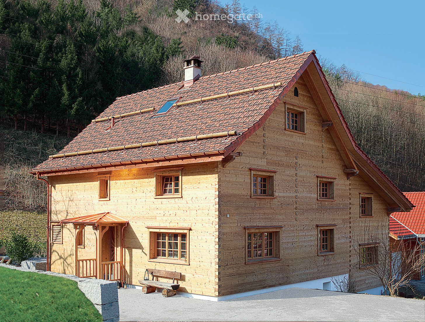 A two-story wooden house with a tiled roof, multiple windows, and a small porch with a bench in front.