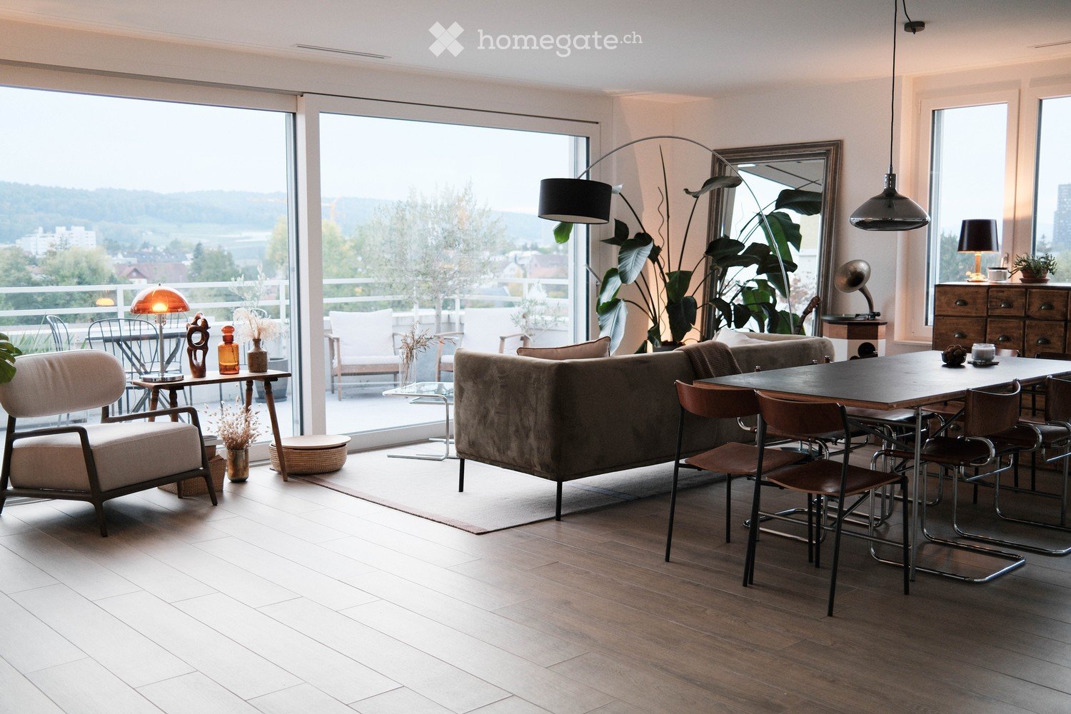 Modern living room with large windows offering a scenic view, featuring a sofa, armchair, dining table with chairs, floor lamp, and decorative items.