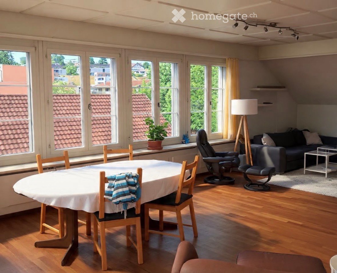 Bright living and dining area with large windows, wooden flooring, a round dining table with chairs, a sofa, a coffee table, a floor lamp, and a potted plant on the windowsill.