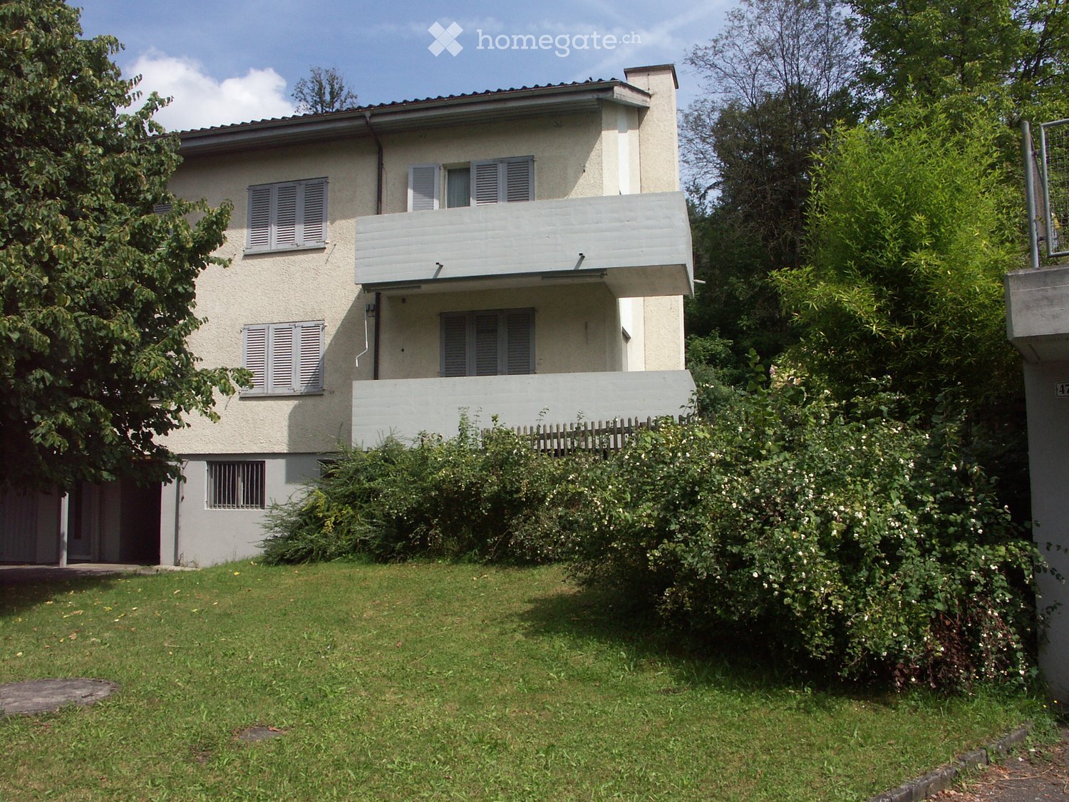 Two-story house with light-colored walls, multiple windows with shutters, a small balcony on the upper floor, and a well-maintained garden with greenery and a wooden fence.