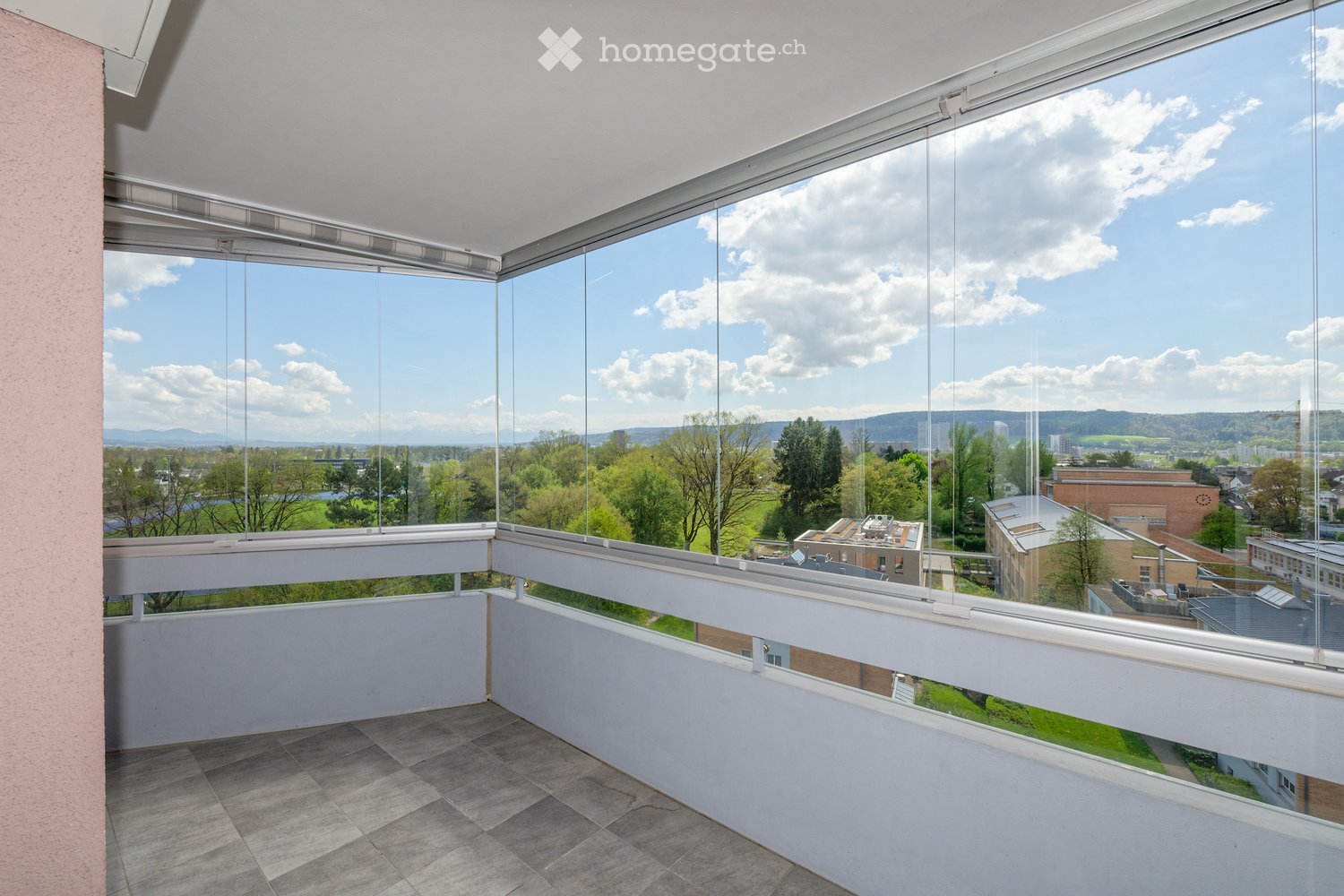Modern balcony with large glass windows offering a scenic view of a green landscape and distant buildings.