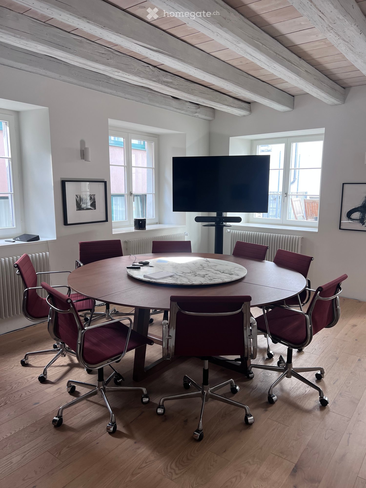Bright room with wooden floors, large windows, round table with marble top, six chairs, and a large screen on a stand.