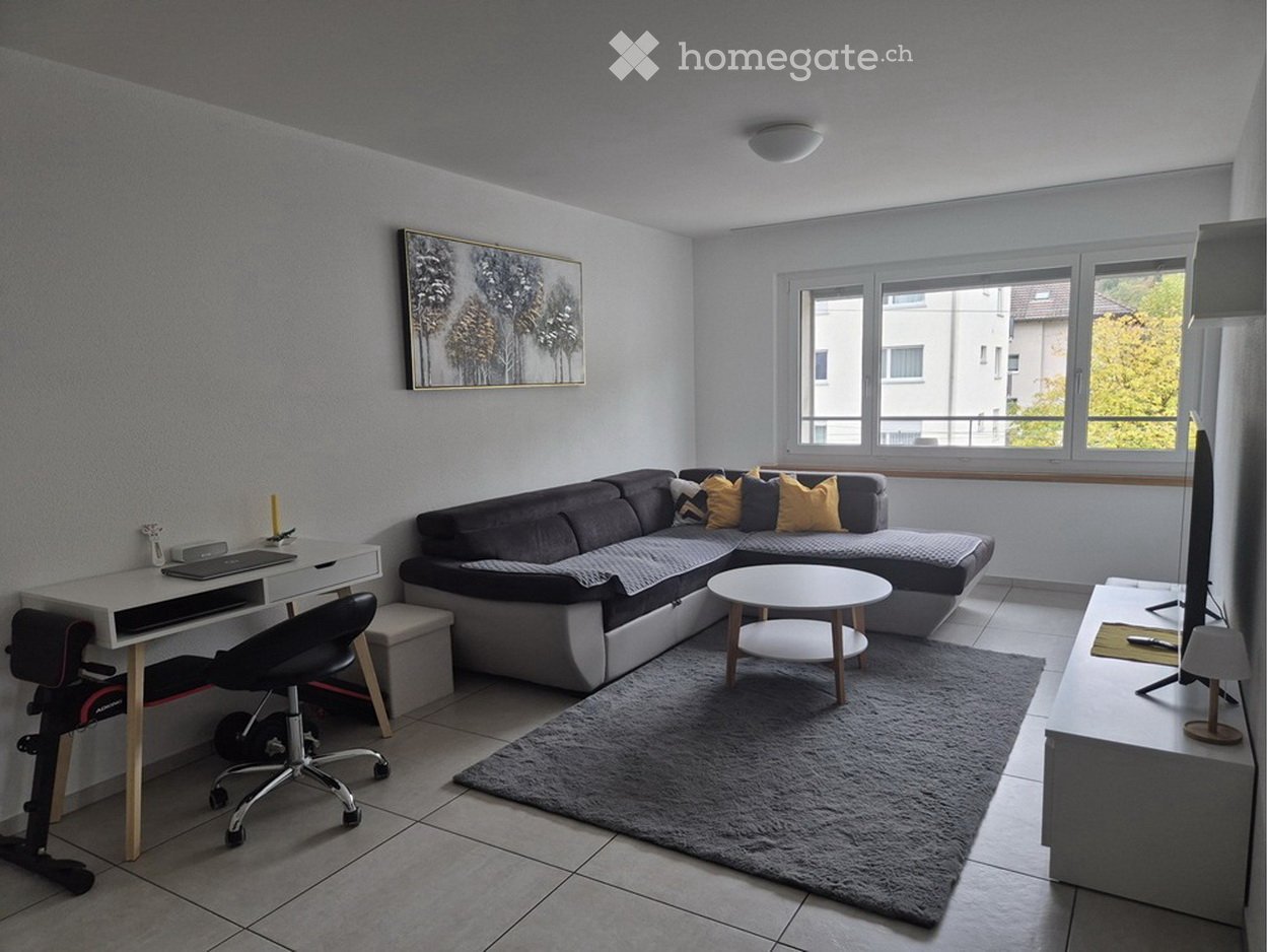 Modern living room with a grey sectional sofa, coffee table, desk with laptop, chair, and a TV screen. Large windows provide natural light.