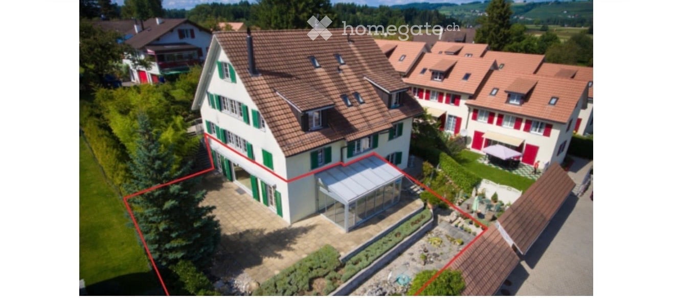 Aerial view of a two-story house with a red-tiled roof, green shutters, and a modern glass extension. The house is surrounded by a well-maintained garden and is part of a residential neighborhood with similar houses in the background.