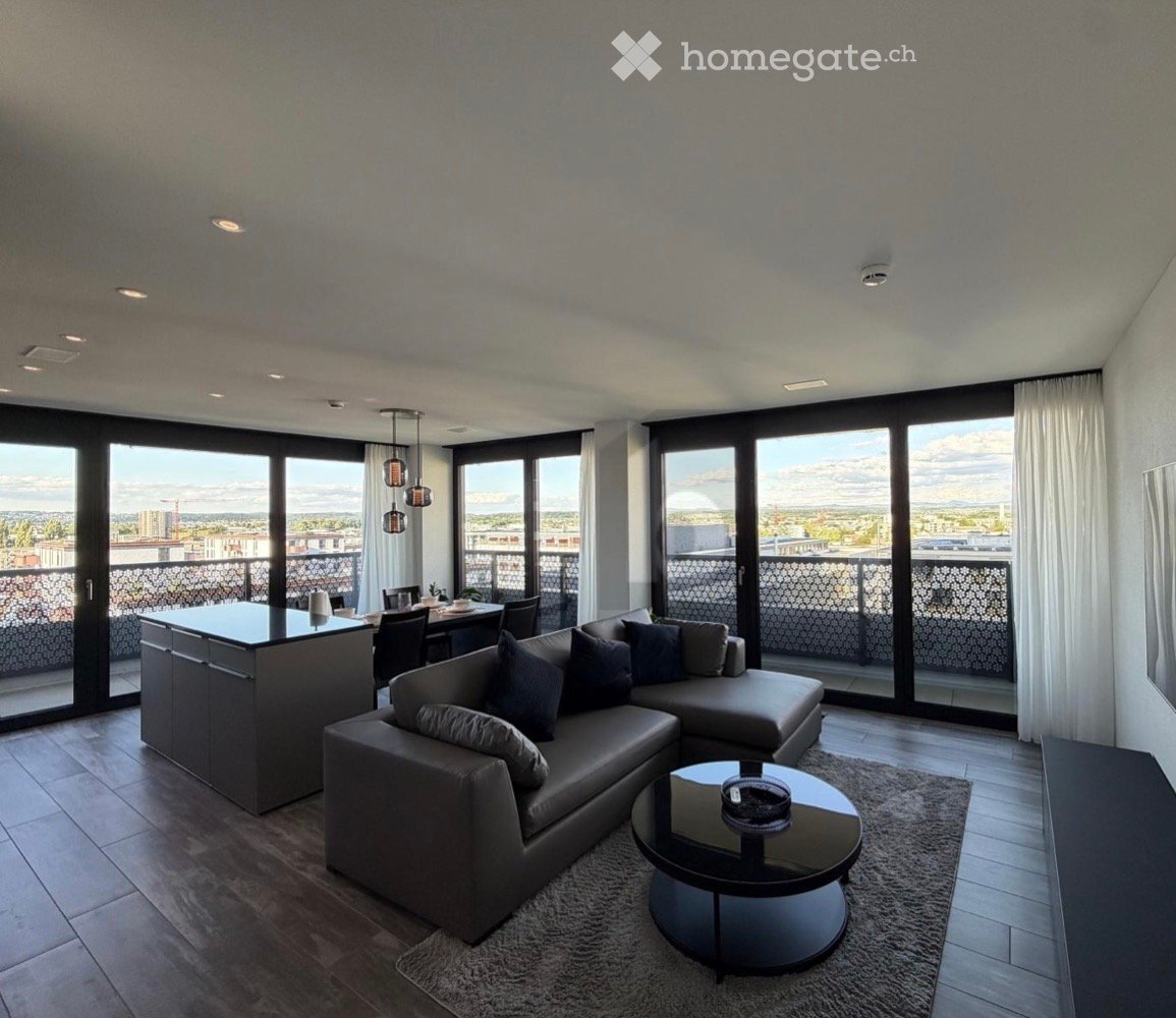 Modern living and dining area with large windows offering a city view, featuring a grey sectional sofa, coffee table, dining table with chairs, and a kitchen island.