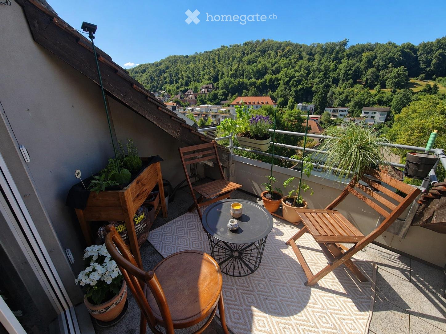 Balcony with wooden chairs, a small round table, and potted plants, offering a scenic view of a green, hilly landscape.