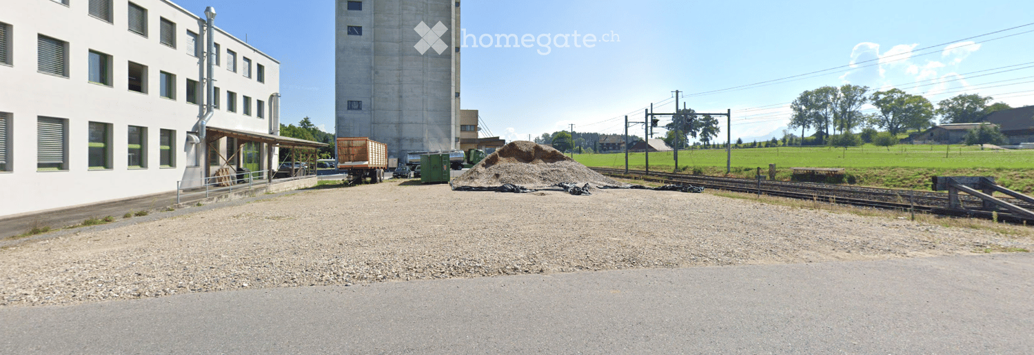 Industrial building with a tall silo, gravel area, and railway tracks in a rural setting.