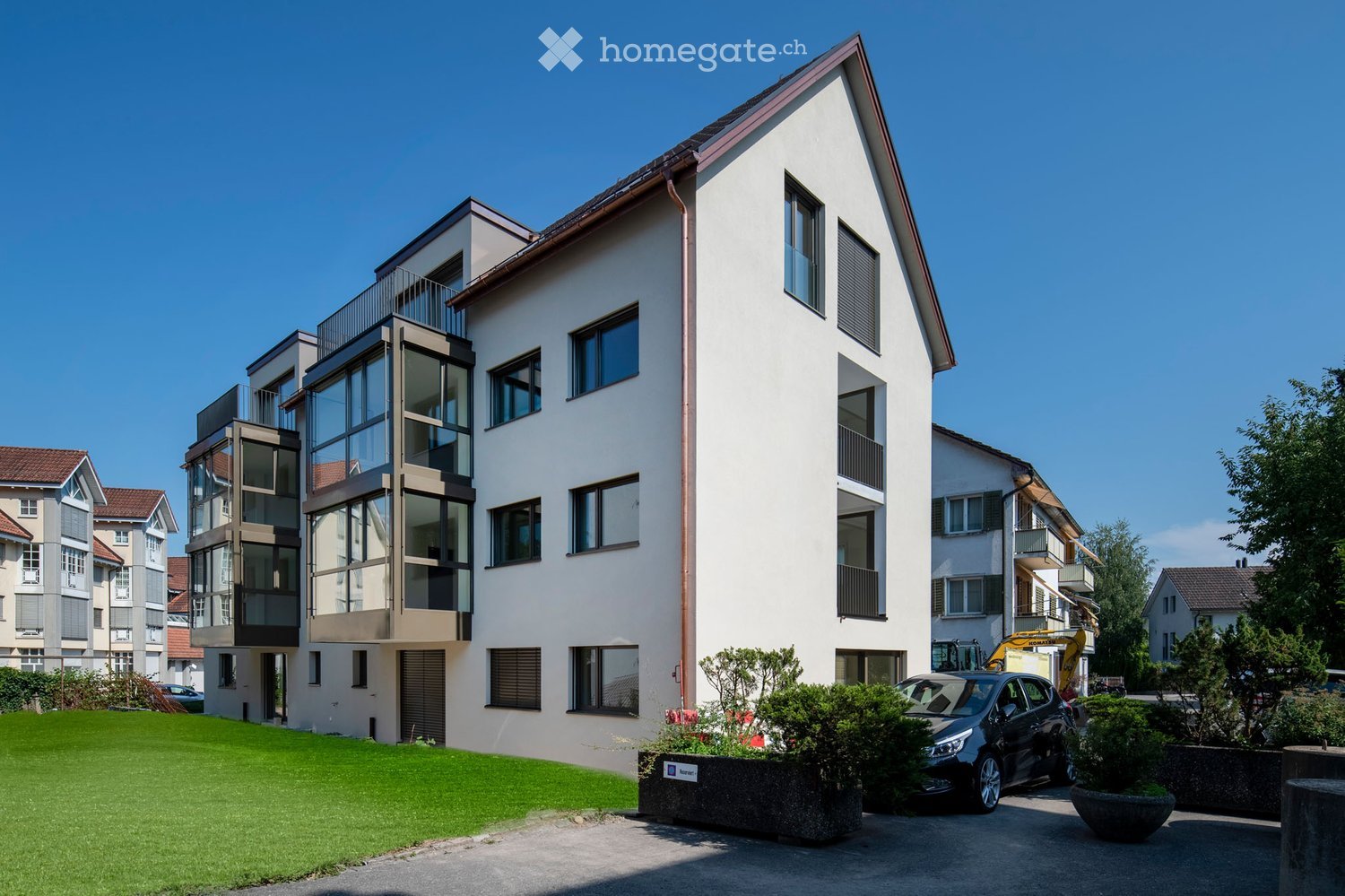 Modern multi-story residential building with white facade, large windows, and balconies. Features a well-maintained lawn, parked car, and surrounding greenery.