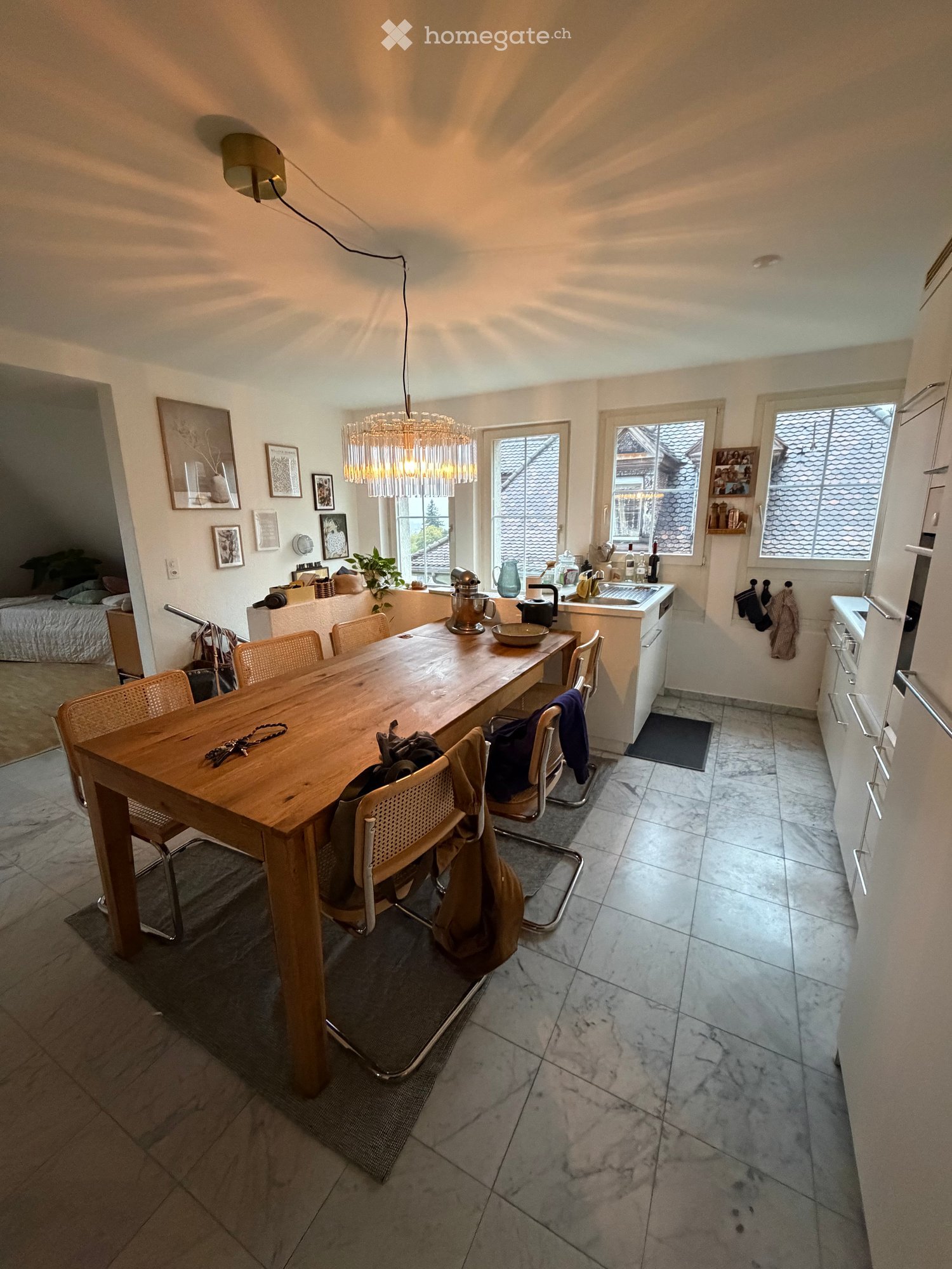 Bright kitchen with a wooden dining table, chairs, sink, and various kitchen items. The room features large windows, a chandelier, and a view of the roof from outside.