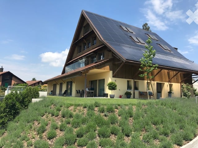 Modern two-story house with a steeply pitched roof, large windows, and a spacious balcony area. The exterior features light yellow walls, wooden accents, and a well-maintained lawn with shrubs and small trees.