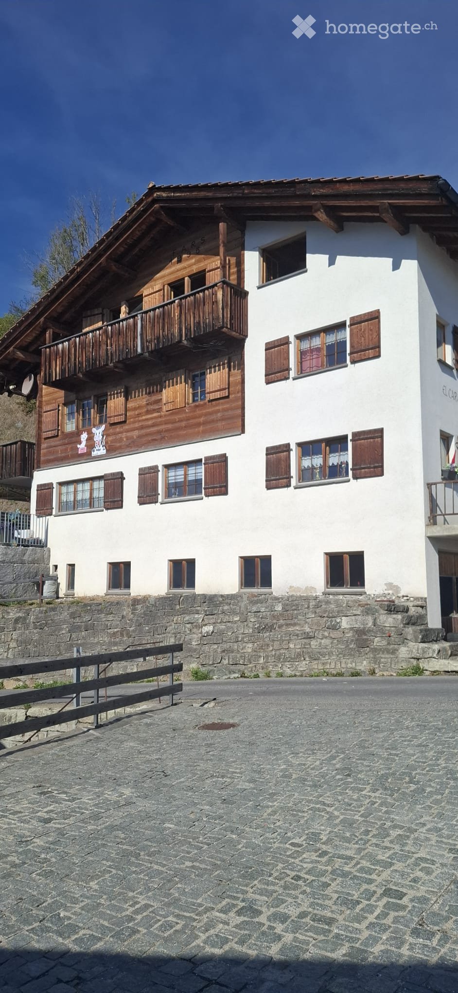 Two-story traditional house with white walls, wooden balconies, and shutters. The ground floor has a stone base and a wooden fence in front.