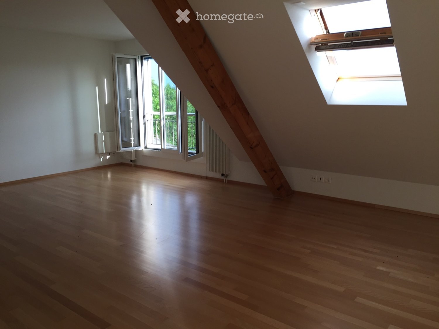 Empty attic room with wooden flooring, large windows, and skylight, featuring exposed wooden beams and radiators.