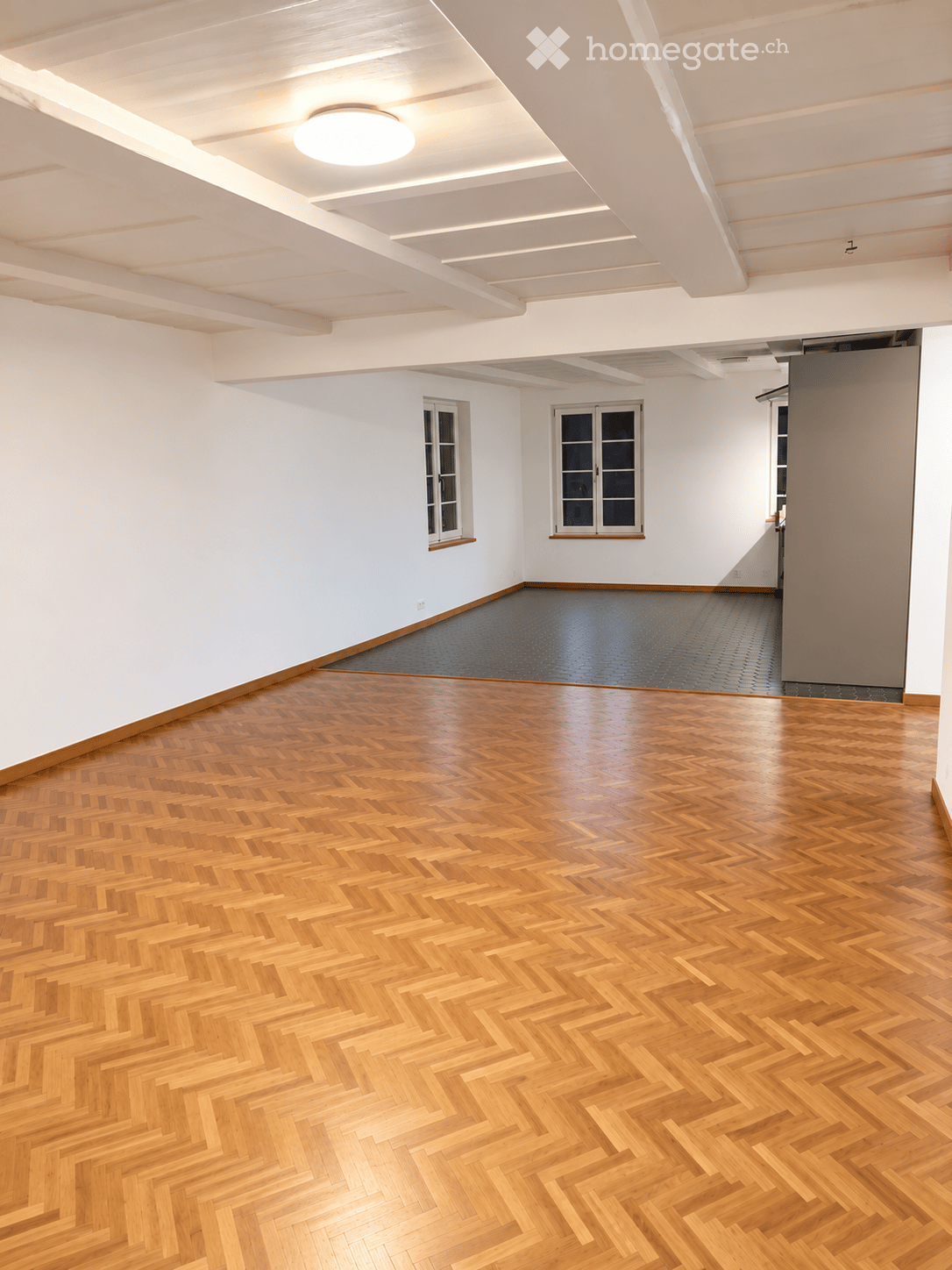 An empty room with polished wooden herringbone flooring, white walls, and large windows. The ceiling features recessed lighting and exposed beams.