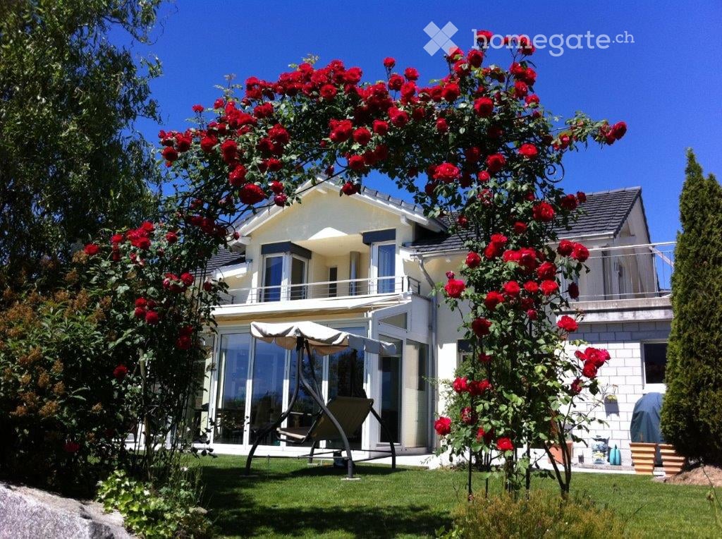 A modern two-story house with a balcony, surrounded by a well-maintained garden featuring red roses and a swing chair.
