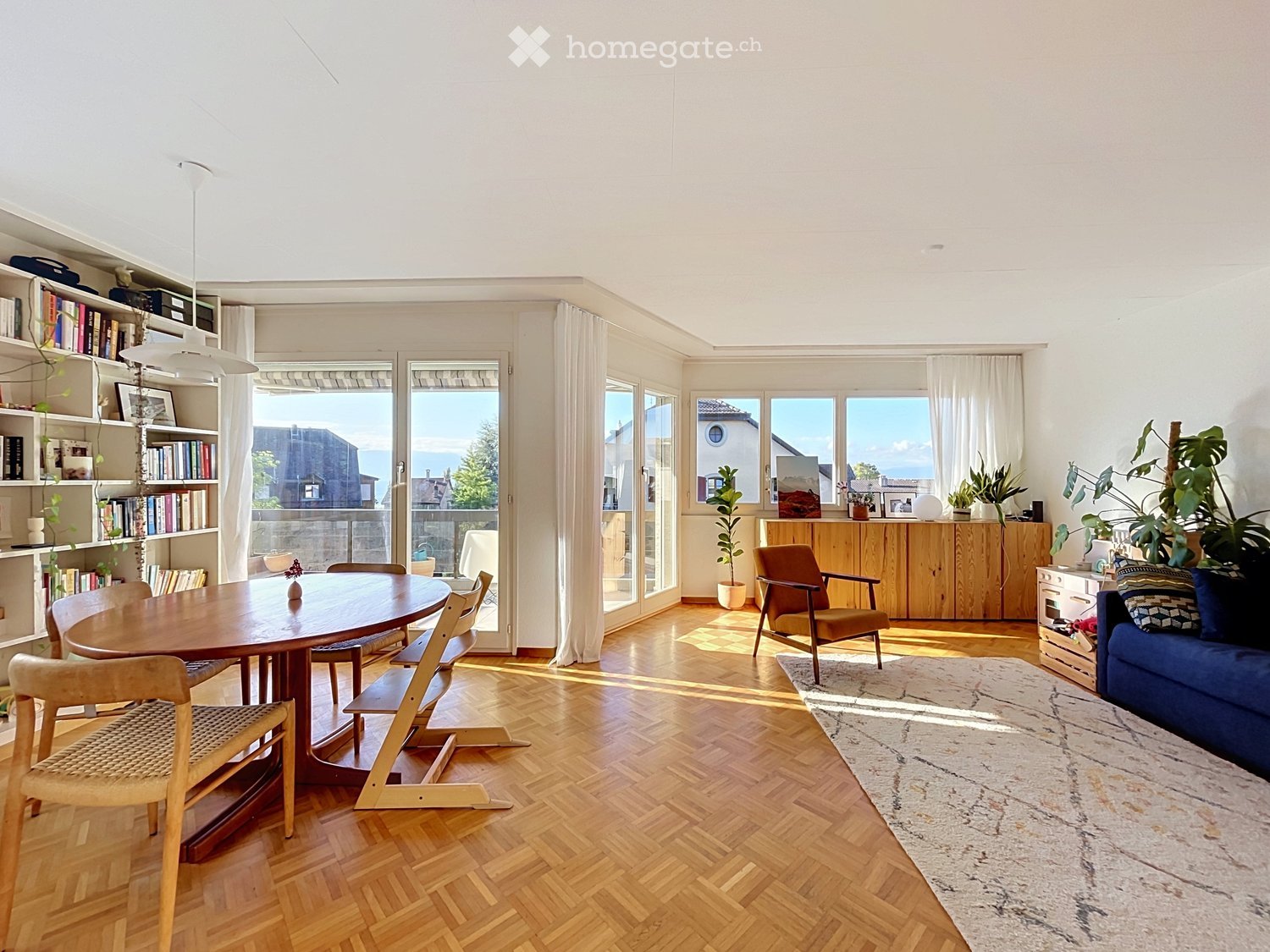 Bright, open living and dining area with wooden flooring, large windows, and a view. Features a blue sofa, wooden dining table with chairs, bookshelf, and potted plants.