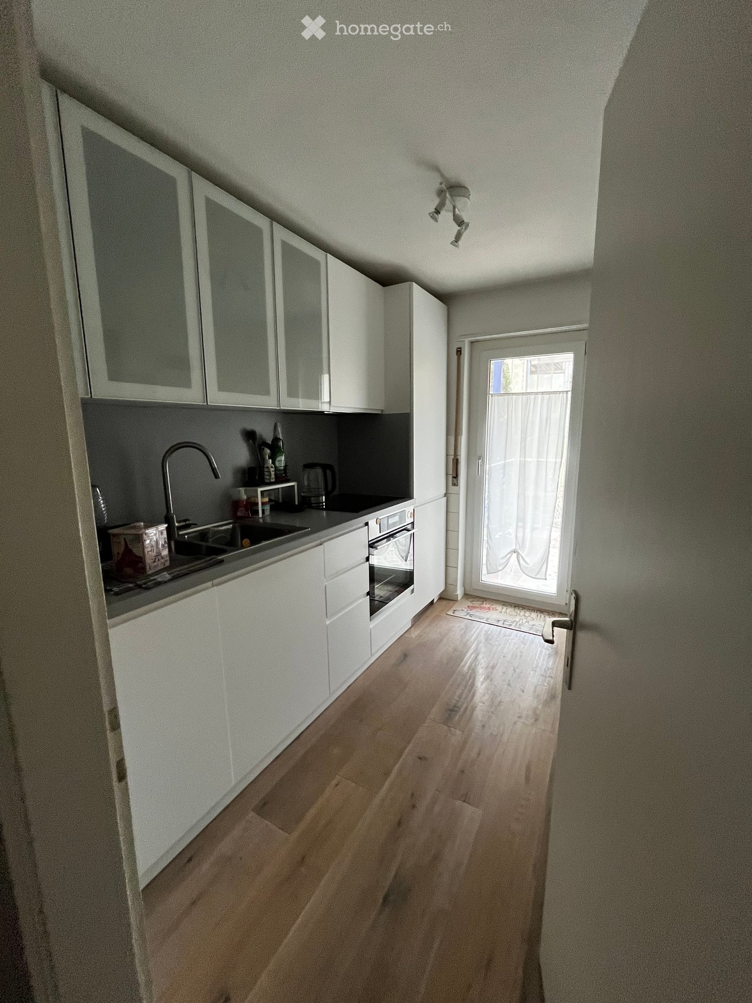 Modern kitchen with white cabinets, wooden flooring, and a sink with a faucet. A small window with a curtain is visible, and the space is well-lit with ceiling lights.