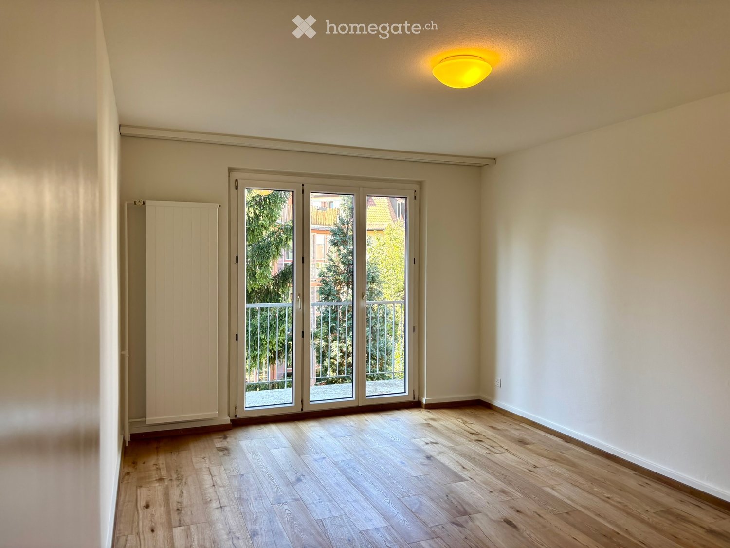 Empty room with wooden flooring, white walls, large sliding glass doors leading to a balcony, and a radiator on the left side.