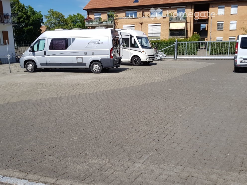 Parking area with two vans and a white van, surrounded by a modern apartment building with balconies and greenery.
