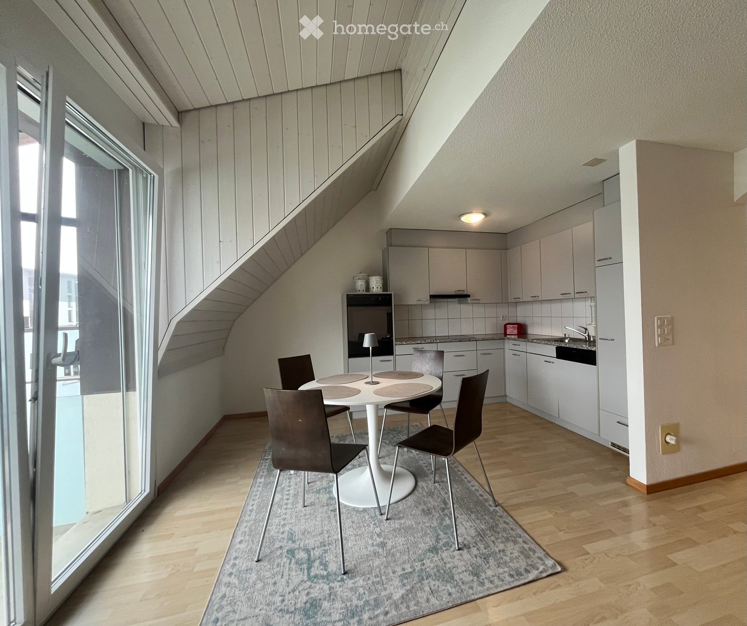 A modern, compact kitchen and dining area with white cabinets, a round white dining table, four black chairs, and a small oven. The space features wooden flooring and large windows letting in natural light.