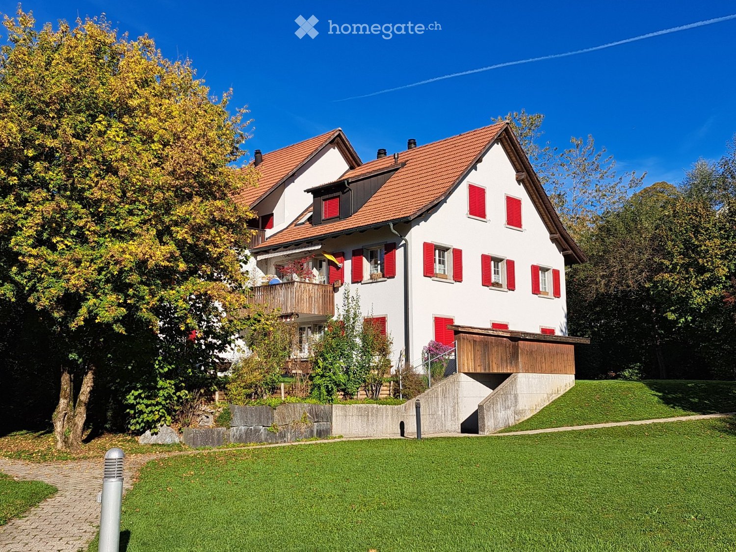 Two-story house with red shutters, red-tiled roof, and a raised ground floor. The house features a wooden balcony and is surrounded by a well-maintained lawn and trees.
