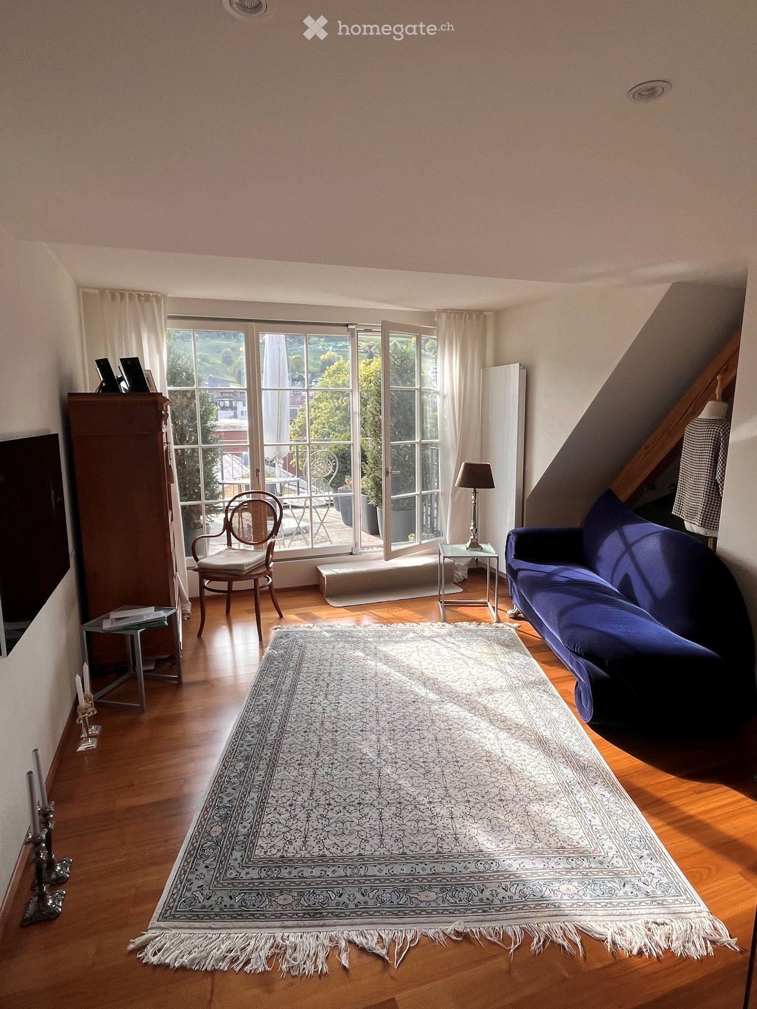 Bright living room with wooden flooring, large windows opening to a balcony, a blue sofa, a patterned rug, a wooden chair, a small table with a lamp, and a cabinet with books and decorative items.