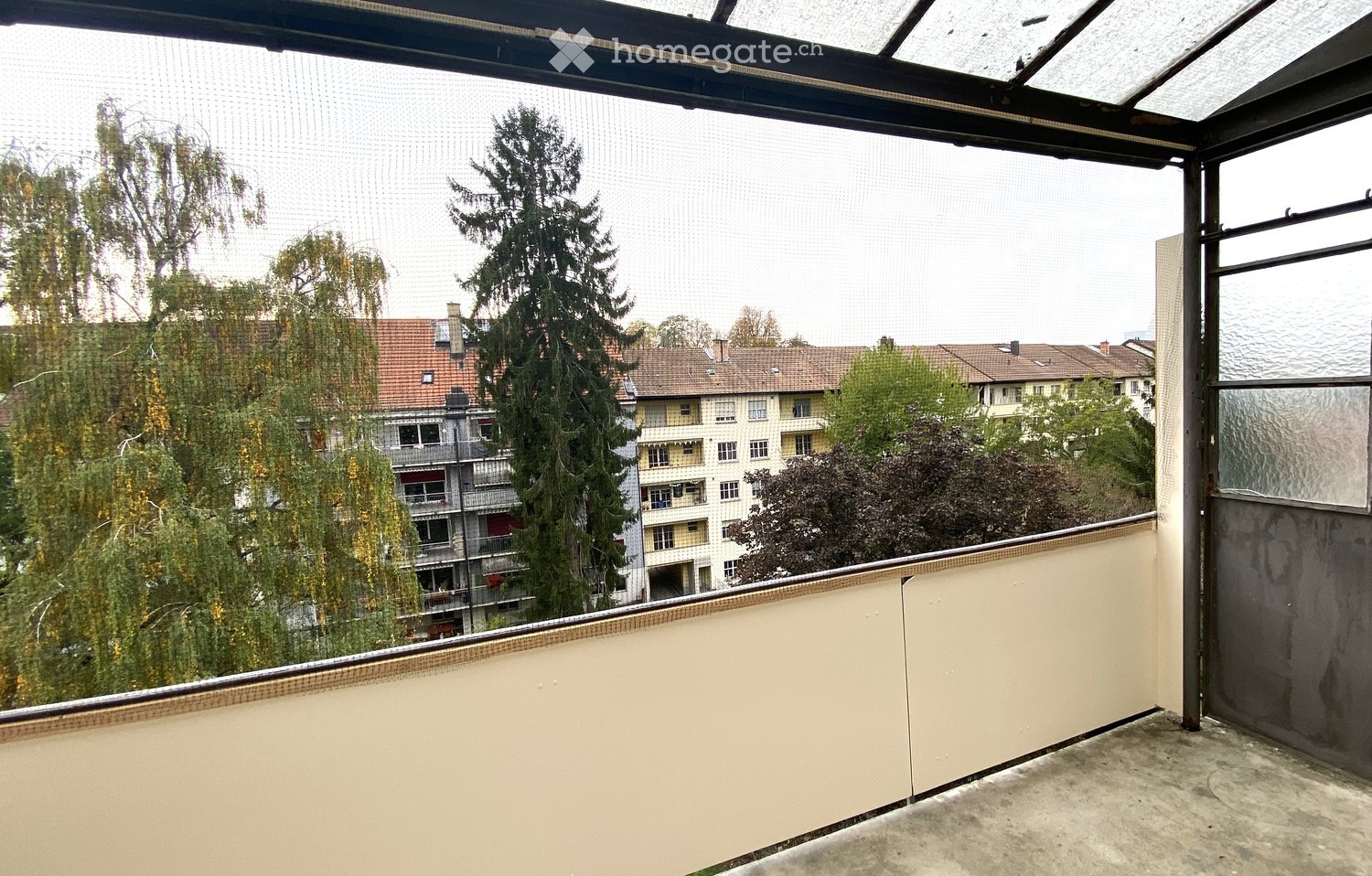Balcony with glass enclosure, overlooking residential buildings and trees.