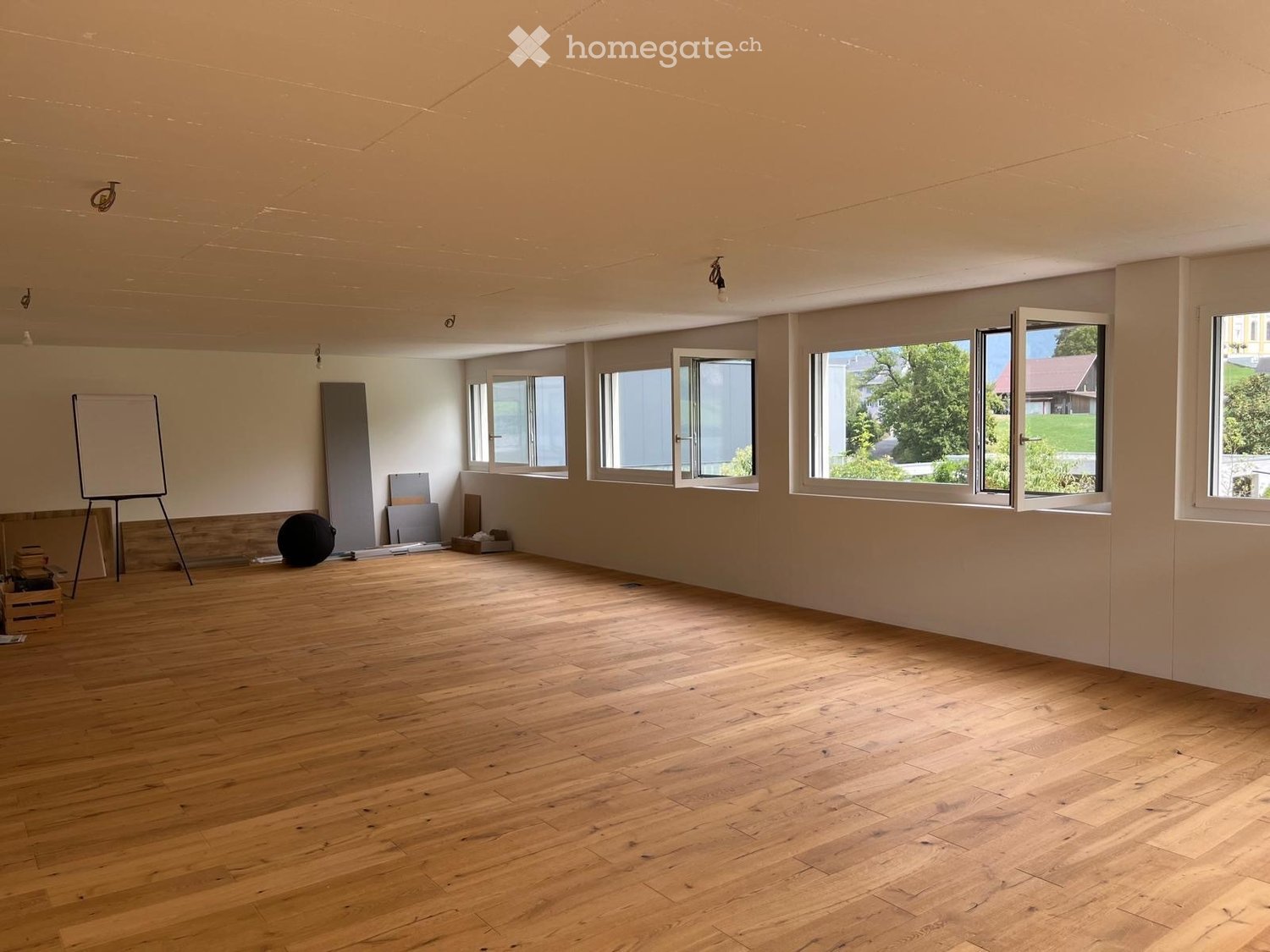 Empty room with wooden flooring, large windows, and minimal furnishings including a whiteboard, a ball, and some stacked items.