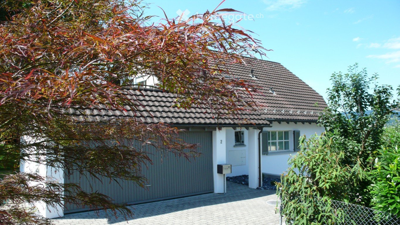 Single-story house with a tiled roof, white exterior, and a grey garage door. Surrounded by trees and greenery.