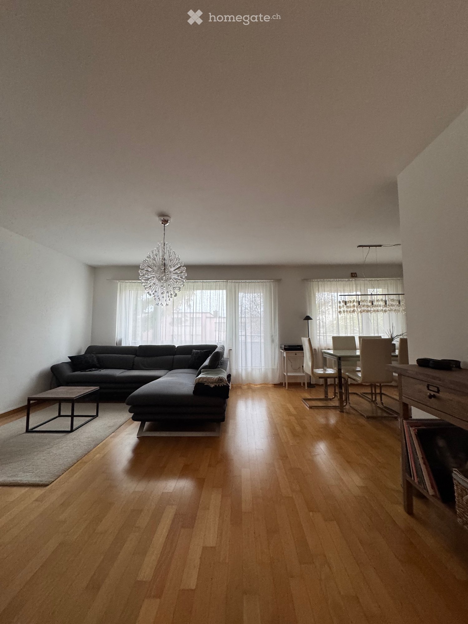 Open-plan living and dining area with wooden flooring, a sectional sofa, coffee table, dining table with chairs, and modern lighting fixtures.