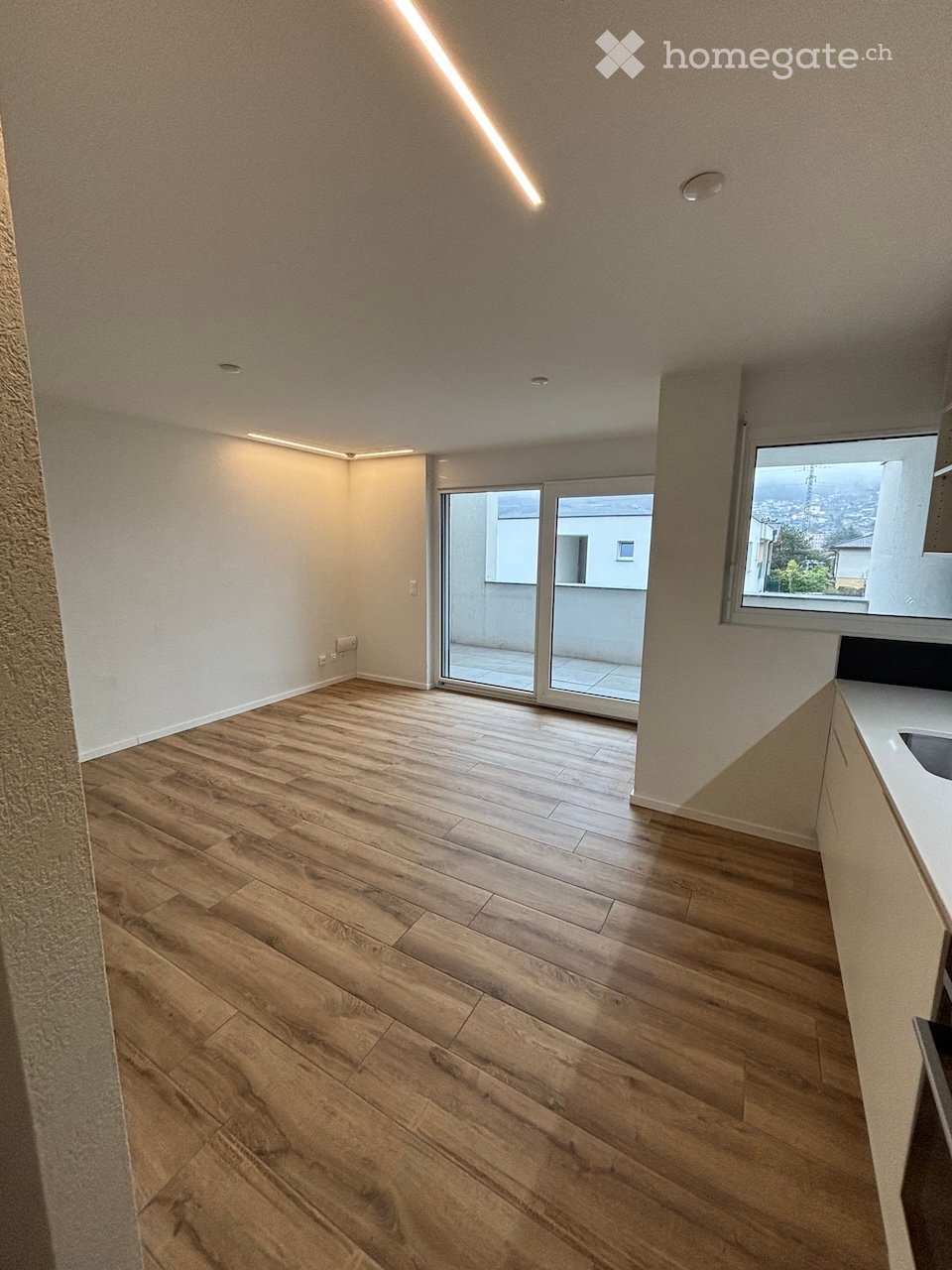 Empty room with wooden flooring, white walls, and a large window with a view. A sliding glass door leads to a balcony.