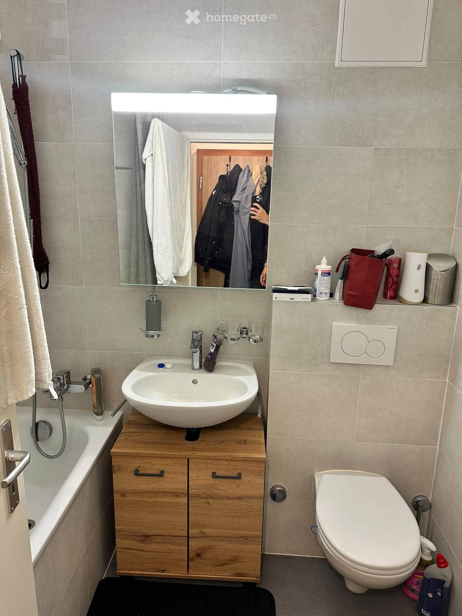 Modern bathroom with a white sink on a wooden cabinet, a large mirror with built-in lighting, a toilet, a bathtub, and various toiletries on the counter.