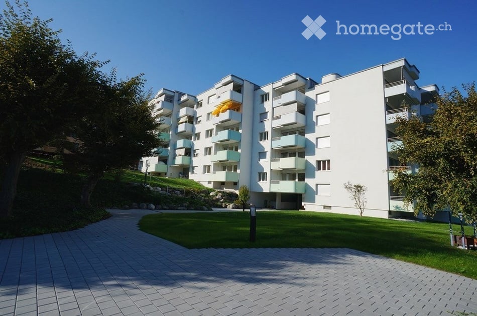Modern multi-story apartment building with white facade, multiple balconies, and surrounding greenery.