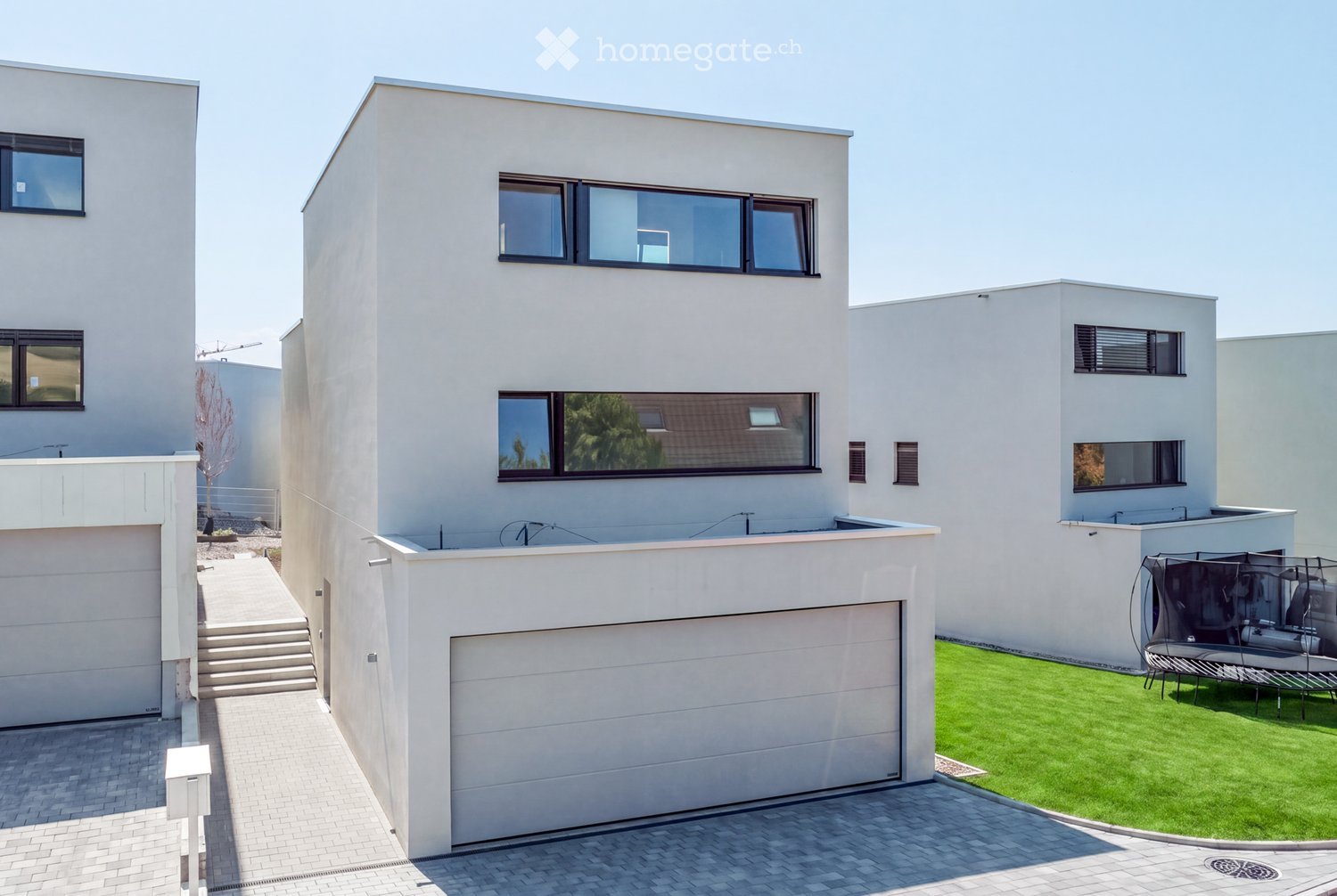 Modern three-story house with white exterior, large windows, garage, and a well-maintained lawn with a trampoline.