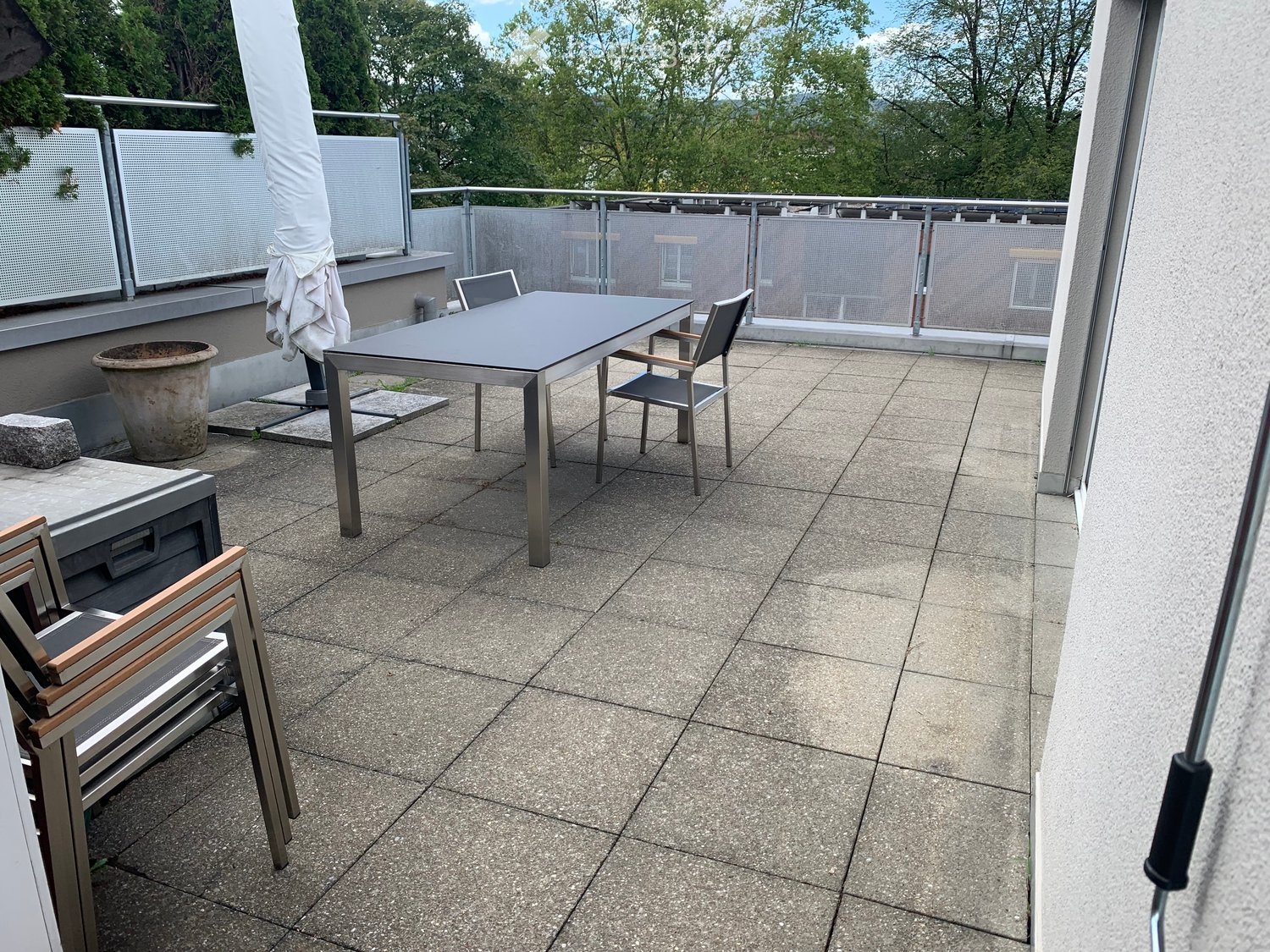 Balcony with tiled floor, metal table, and chairs, featuring a view of greenery and a covered umbrella.