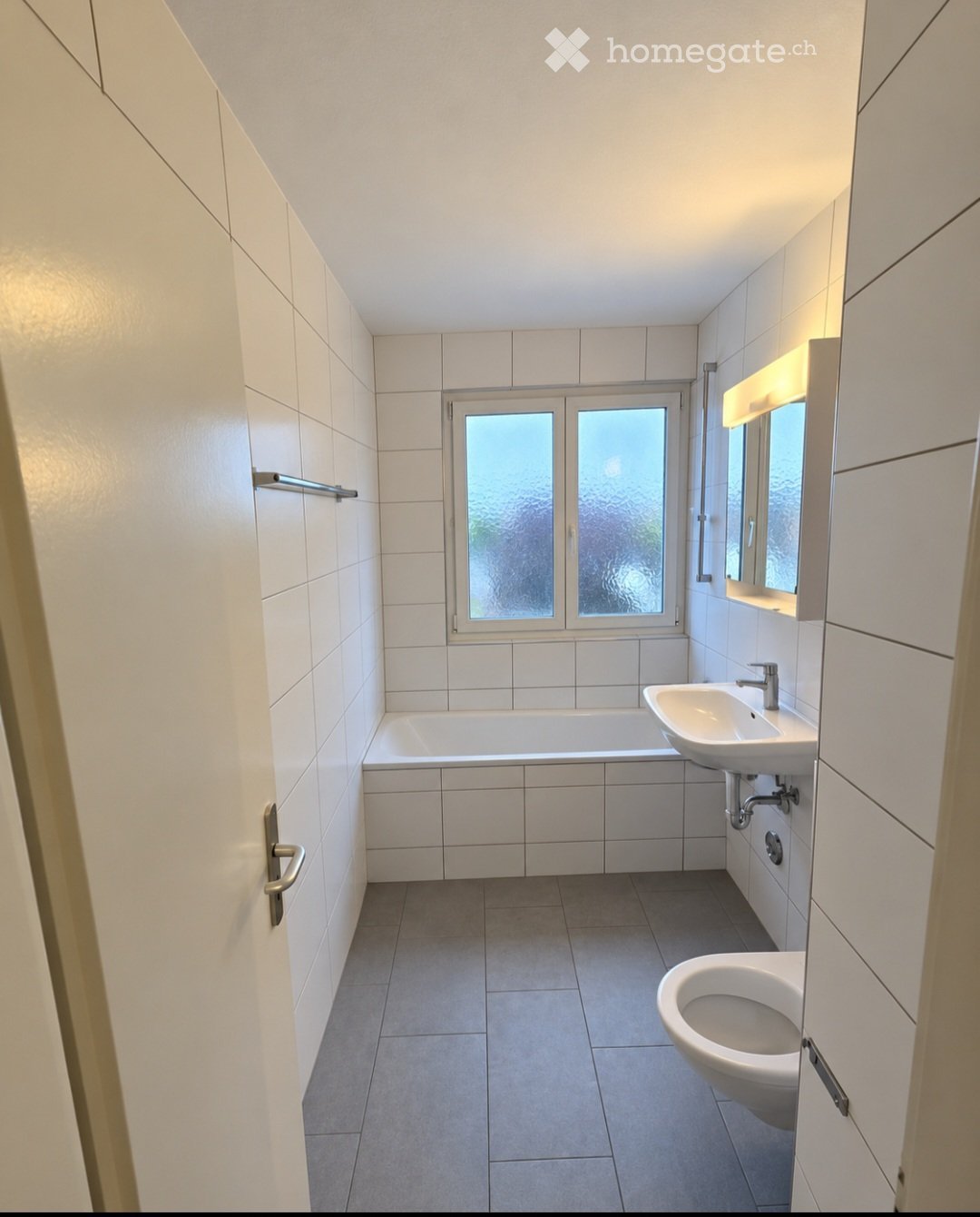 Modern bathroom with white tiled walls and floor, featuring a bathtub, sink, toilet, and a window with frosted glass panes.