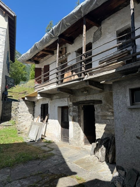 A rustic stone building with a wooden balcony and weathered exterior, situated in a mountainous area with a clear blue sky in the background.