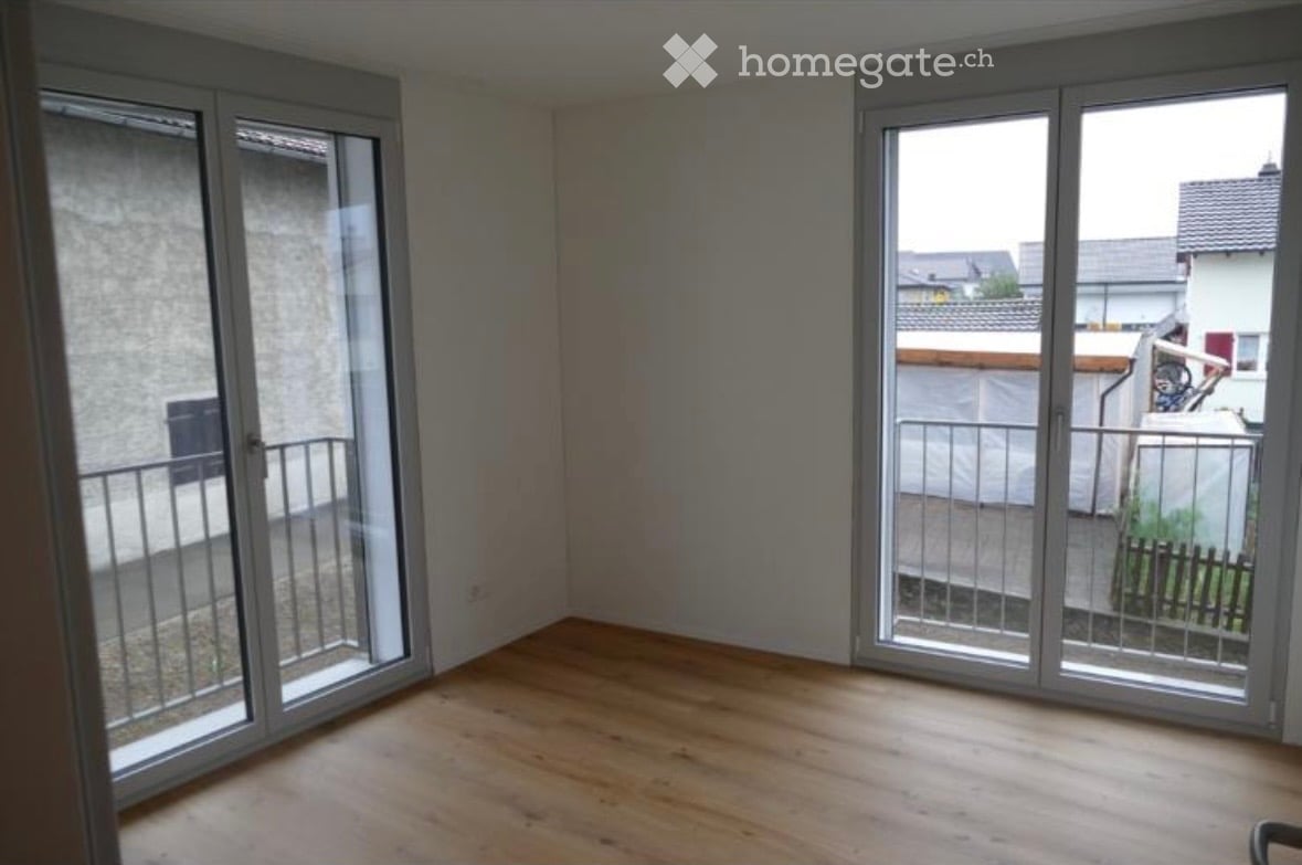 Empty room with wooden flooring, large sliding glass doors leading to a balcony, and a view of neighboring buildings.