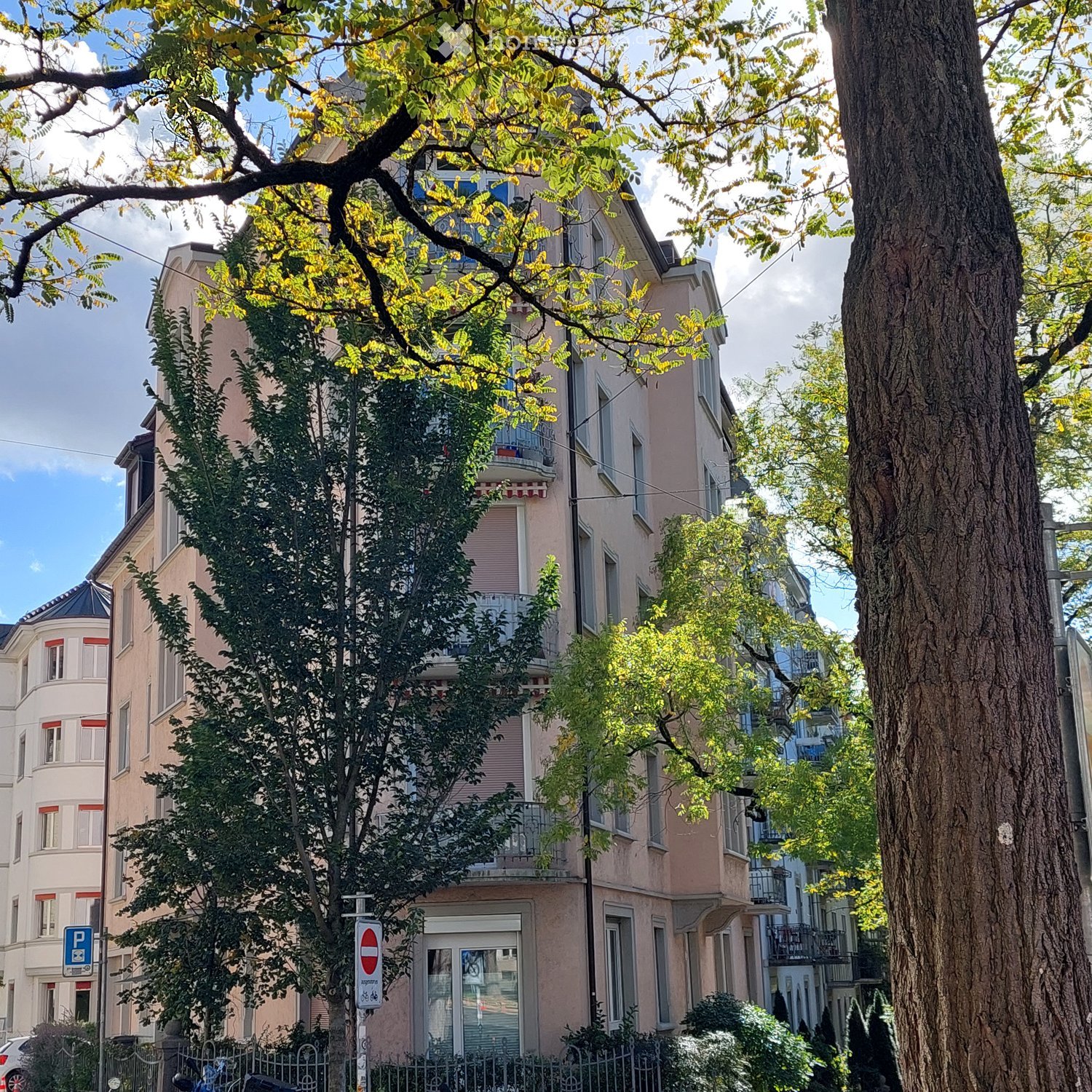 Multi-story residential building with balconies, surrounded by trees and greenery.