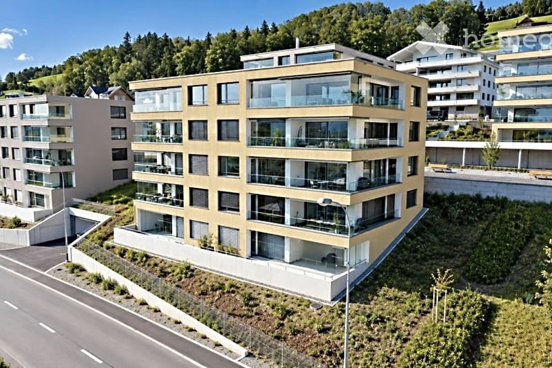 Modern multi-story residential building with balconies and large windows, surrounded by greenery and other buildings.